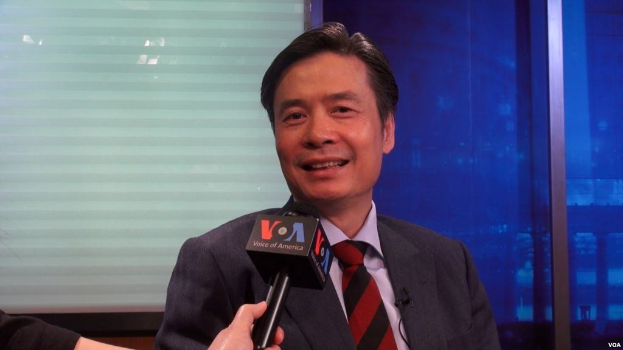 Ein Mann in Anzug und Krawatte hält ein Mikrofon und hat eine Hand auf der linken Seite sichtbar. Im Hintergrund ist ein Fenster und eine Wand zu sehen.