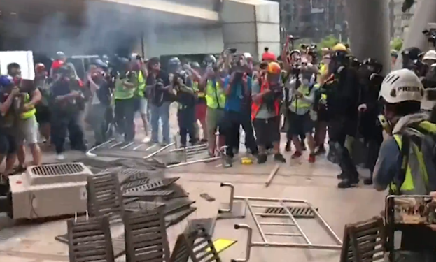 Eine Gruppe von Menschen steht vor einem rauchenden Gebäude, einige tragen Helme und Jacken, andere halten Kameras, mit verstreuten Metallobjekten und Gegenständen auf dem Boden, vor einer Kulisse aus Stadtpfeilern und Gebäuden.