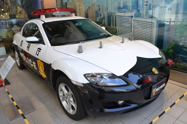 Polizeiauto vor einer Glaswand geparkt, mit sichtbarem Rad und einer Texttafel daneben, die Gebäude, Bäume und Himmel durch die Glaswand zeigt.