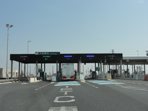 Mautstelle mit einem vor ihr geparkten Lkw auf einer Straße mit weißen und blauen Markierungen, Laternen, Schildern und anderen Gegenständen, mit Gebäuden, Bäumen und einem klaren blauen Himmel im Hintergrund.