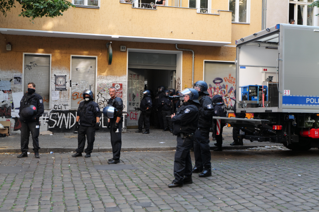 Eine Gruppe von Polizisten in Helmen und mit Waffen steht vor einem Gebäude mit Fenstern, Rollläden und Graffiti, daneben parkt ein Lastwagen und im Hintergrund sind Bäume zu sehen.