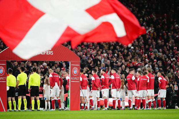 Eine Gruppe von Menschen auf einem Fußballfeld mit einer rot-weißen Fahne im Vordergrund, einem Bogen mit Text "Bayern München vs Bayern München Wett-Tipps & Vorschau" im Hintergrund und einer großen Menge im Stadion.