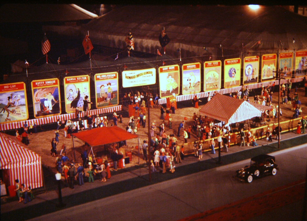 Altes Schwarz-Weiß-Foto eines nächtlichen Volksfestes mit Menschen, einem Auto, Masten mit Fahnen, einem Zaun, einem Zelt, illustrierten Tafeln und einer Straßenlaterne.