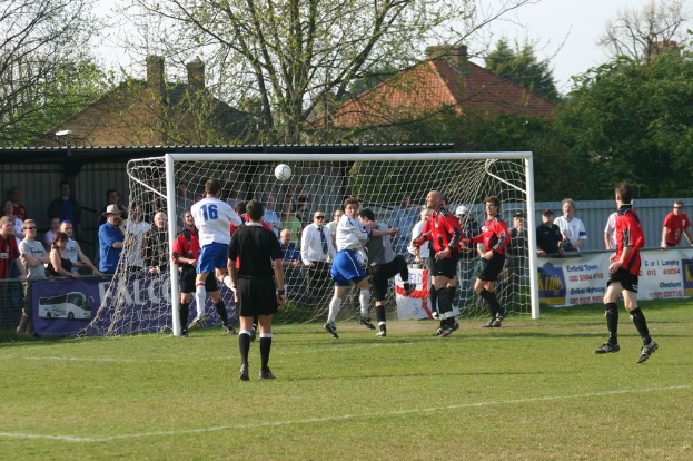 Spieler sind in einem Füßballspiel auf einem Feld mit einem Tornetz beteiligt, während Zuschauer dahinter stehen; Bäume und Häuser sind im Hintergrund sichtbar.