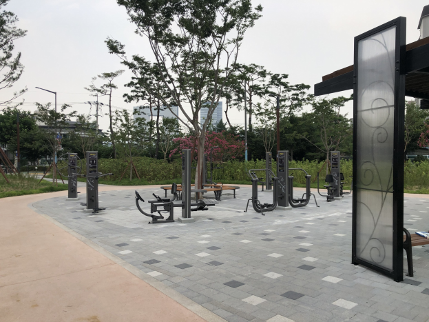 Outdoor-Park mit verschiedenen Fitnessgeräten, Bänken, Bäumen, Pflanzen, Gras, Pfosten, Lichtern, Kabeln, Gebäuden und Himmel als Hintergrund.