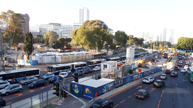 Eine belebte Stadtstraße mit zahlreichen Autos, Bussen, Lastwagen, Verkehrszeichen, Pfosten, Schildern, Bäumen und Gebäuden unter einem klaren blauen Himmel.