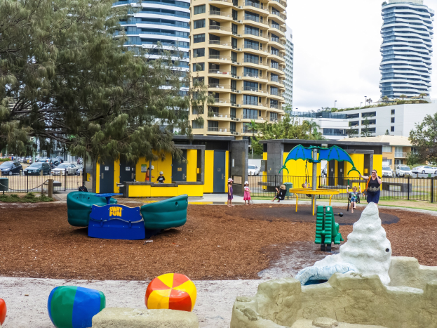 Spielplatz mit Kindern auf Geräten, umgeben von Bäumen und hohen Gebäuden in einer städtischen Umgebung mit Fahrzeugen auf der Straße und sichtbarem Himmel oben.