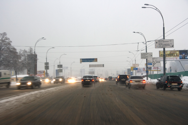 Eine belebte Stadtstraße mit Fahrzeugen auf einer schneebedeckten Straße, gesäumt von Laternen, Texttafeln, Bäumen und Gebäuden unter einem verschneiten Himmel.