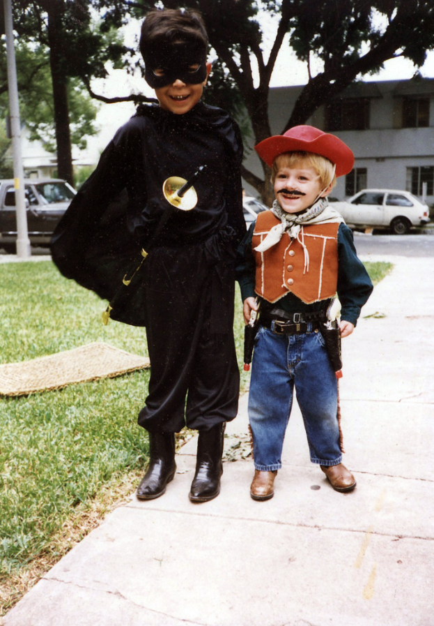 Kinder in verschiedenen Kostümen stehen auf einer Straße mit Gebäuden, Bäumen und Fahrzeugen im Hintergrund.