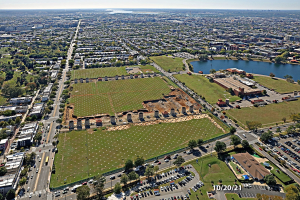Luftaufnahme einer Stadt mit Gebäuden, Bäumen, Gras, Straßen mit Fahrzeugen, Wasser und Wolken, die einen großen zentralen Bereich für einen Park zeigt.