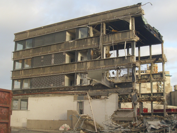 Ein teilweise abgerissenes Gebäude inmitten einer Straße, mit einem Container auf der linken Seite und einem bewölkten Himmel im Hintergrund, das Anzeichen von Verfall mit zerbrochenen Fenstern und verstreuter Trümmern zeigt.