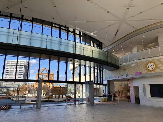 Innenansicht eines großen Gebäudes mit zahlreichen Fenstern, Deckenleuchten, einer Wanduhr, einem Fernseher, einem Anzeigebildschirm, Holzkonstruktionen, einem Holzzaun, benachbarten Gebäuden, Bäumen und einem bewölkten Himmel, das als Eingang zu einem neuen Kinderspielplatz dient.