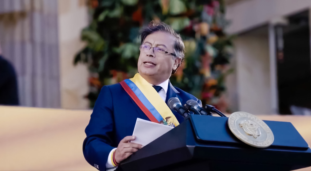 Ein Mann in einem blauen Anzug und Krawatte steht an einem Rednerpult, hält Papiere und spricht in ein Mikrofon, mit einer Pflanze und einer Wand im Hintergrund, was auf eine formelle Umgebung hinweist.