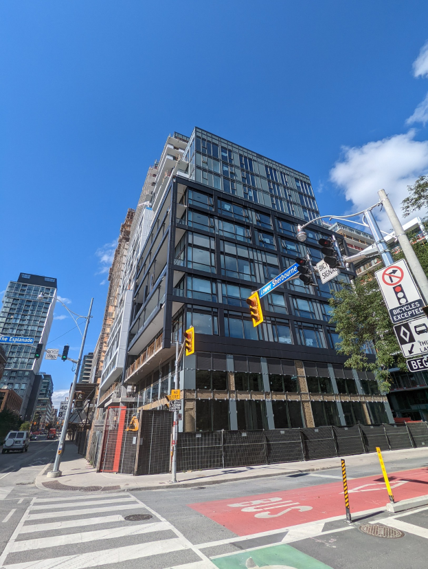 Eine belebte Stadtstraße mit einem hohen Apartment/Wohnhausgebäude zum Verkauf an der 541 East 57th Street, Unit 541 im Hintergrund, mit Fahrzeugen, Verkehrsampeln, Schildern, Bäumen, einem Zaun und einem bewölkten Himmel.