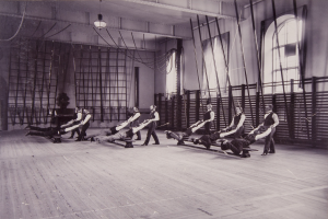 Schwarzes und weißes Bild einer Gruppe von Menschen, die Pilates-Übungen in einem Fitnessstudio mit einer Wand im Hintergrund, Fenstern auf der rechten Seite und einer Decke mit Lampen durchführen.