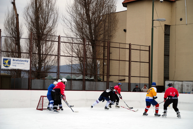 Personen, die Eis-hoc-key auf einem Eisstadion mit Gebäuden, Bäumen, einer Straßenlaterne, einem Namensschild und Zäunen im Hintergrund und einem Himmel spielen.