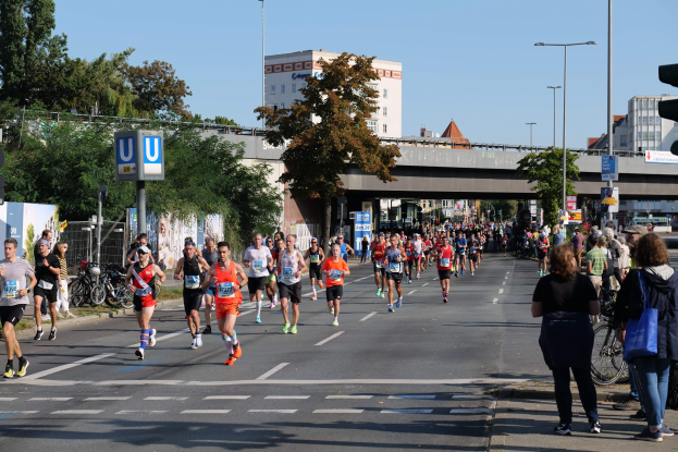 Gruppe von Menschen, die einen Marathon auf einer von Bäumen gesäumten Straße mit Laternen, Schildern, Fahrrädern, einem Zaun, Gras, einer Brücke, Gebäuden und einem klaren blauen Himmel laufen
