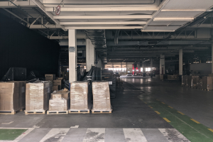 Großer Lagerraum mit gestapelten Kartonagen auf Paletten, beleuchtet von Deckenlampen und gestützt von Säulen, mit einer Deckenanzeige, die ein Flottenmanagementsystem anzeigt.