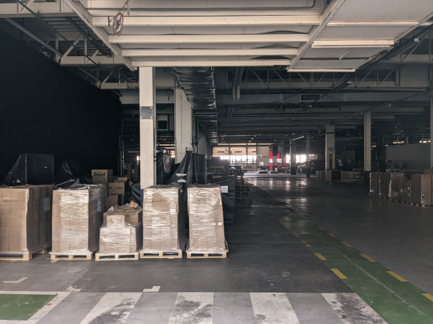 Großer Lagerraum mit gestapelten Kartonagen auf Paletten, beleuchtet von Deckenlampen und gestützt von Säulen, mit einer Deckenanzeige, die ein Flottenmanagementsystem anzeigt.