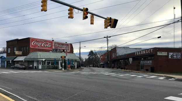 Eine Stadtstraße mit einer Ampel, Fahrzeugen, Gebäuden mit Namensschildern, Strommasten mit Kabeln, Bäumen und einem bewölkten Himmel.