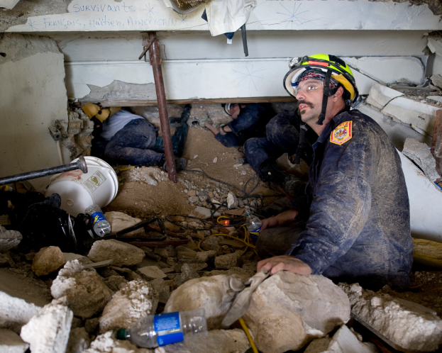 Ein Feuerwehrmann mit Helm sitzt inmitten von Trümmern, darunter Steine und Flaschen, mit einer Wand im Hintergrund, auf der ein Text steht.