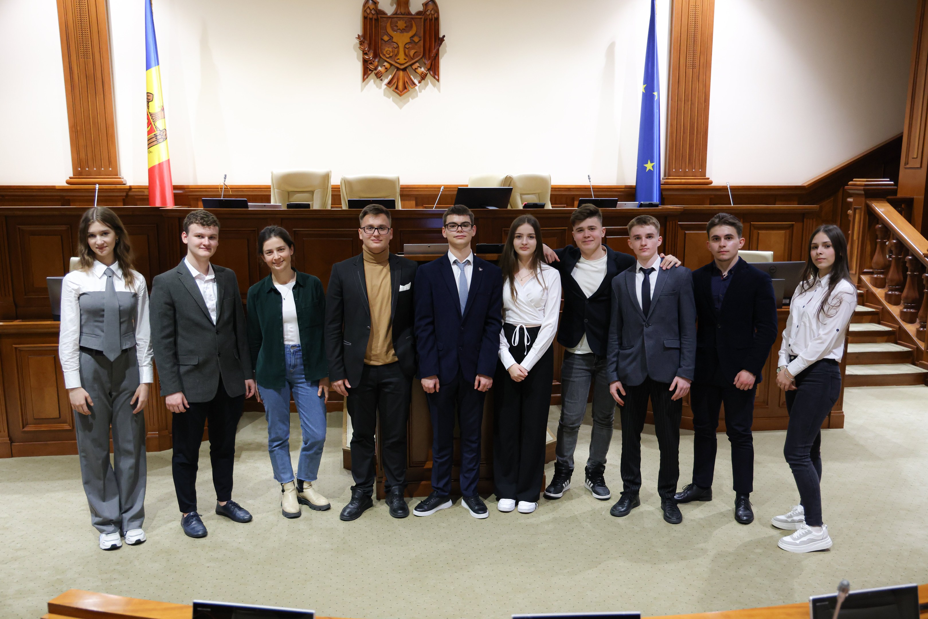 Gruppe von Menschen, die vor einem Gerichtssaal mit Monitoren und Mikrofonen auf Tischen, einer hölzernen Plattform mit Stühlen, Flaggen und einem Wandobjekt im Hintergrund und einer Treppe mit Gel√ändern auf der rechten Seite posieren.