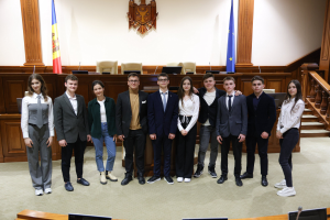 Gruppe von Menschen, die vor einem Gerichtssaal mit Monitoren und Mikrofonen auf Tischen, einer hölzernen Plattform mit Stühlen, Flaggen und einem Wandobjekt im Hintergrund und einer Treppe mit Gel√ändern auf der rechten Seite posieren.