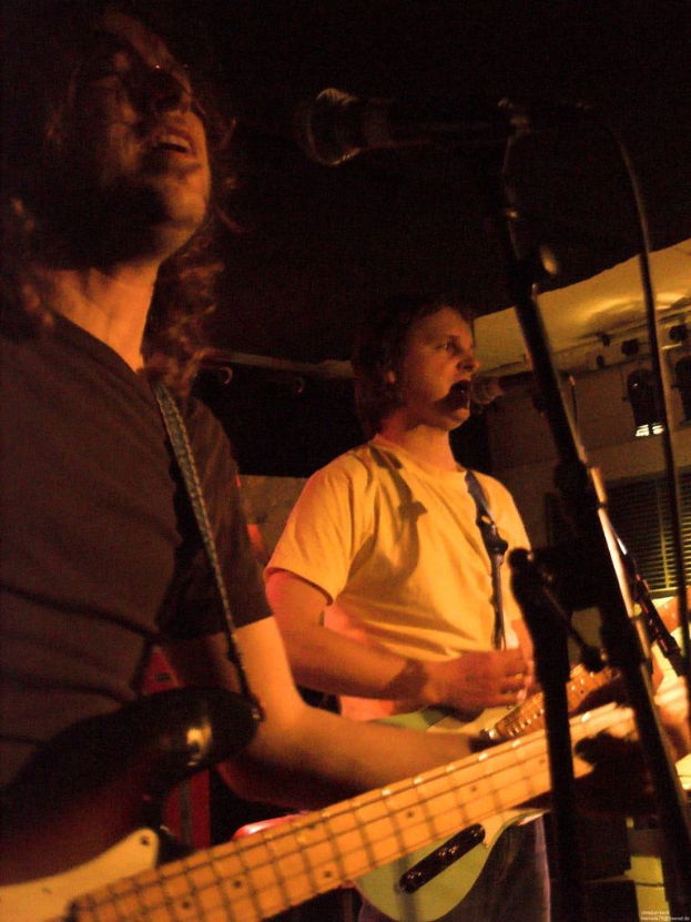 Eine Person in einem weißen Hemd singt in ein Mikrofon und spielt eine grüne Gitarre, während sie neben einer anderen Person in einem schwarzen Hemd und Brille steht, die ebenfalls singt und eine schwarze Gitarre spielt, mit Bühnenlicht und anderen Instrumenten im Hintergrund.