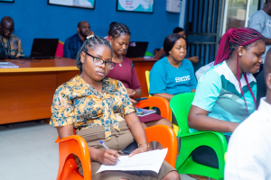 Gruppe von Menschen, die in einem Raum mit Laptops, Büchern und anderen Gegenständen auf Tischen sitzen, gerahmte Gegenstände an der Wand und eine Tür im Hintergrund.