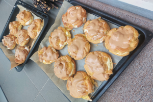Ein Tablett voller gemischter dänischer Teilchen mit einem hellen goldbraunen Farbton und einer fluffigen Textur, einige mit Puderzucker bestäubt, steht auf einer Theke neben einem offenen Fenster und wird von warmem Licht beschienen.