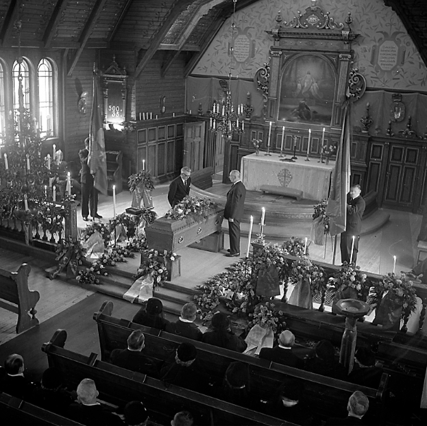 Ein Schwarz-Weiß-Foto von einer Trauerfeier in einer Kirche, mit Sitzenden und Stehenden, Kerzen, Blumensträußen, Fahnen, einem Tisch, einem wandmontierten Rahmen und Fenstern im Hintergrund.